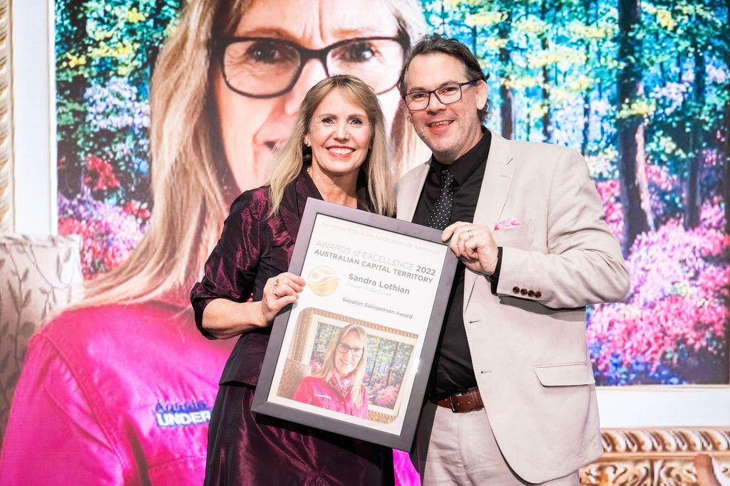 Woman and man holding a framed award, smiling, in front of a backdrop with a woman's image.