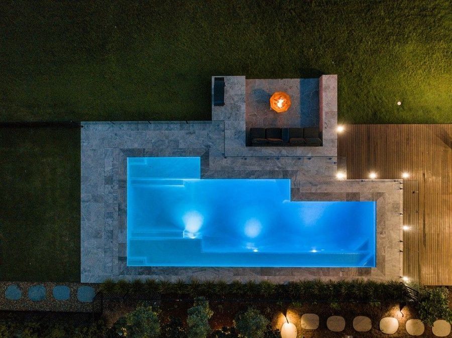 Overhead view of a modern backyard at night with a blue-lit pool, seating area with fire pit, and wooden deck.