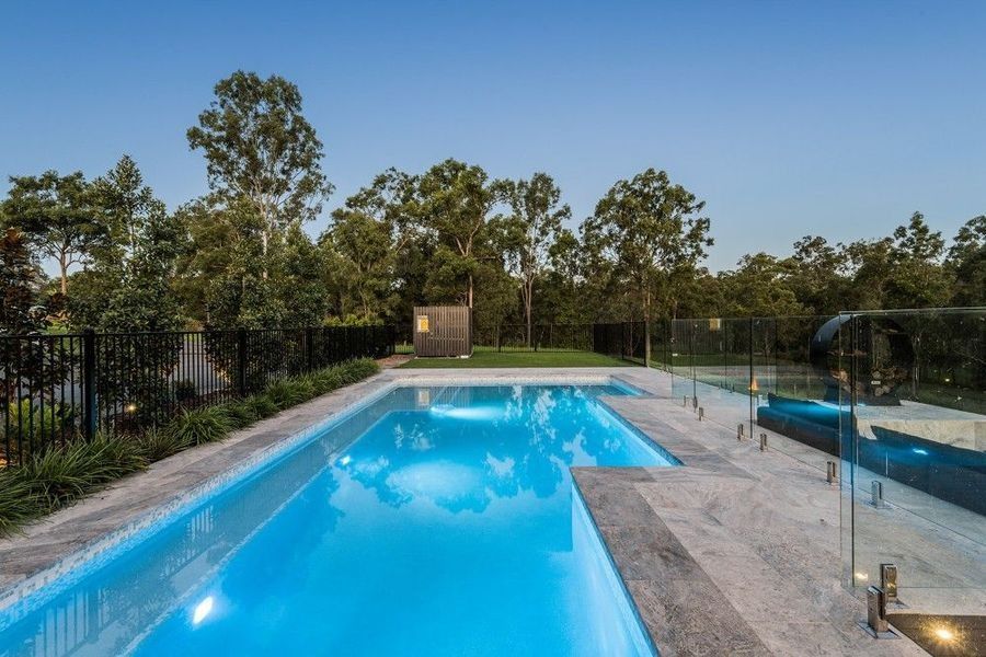 Swimming pool surrounded by gray stone, glass fence, and trees under a blue sky.