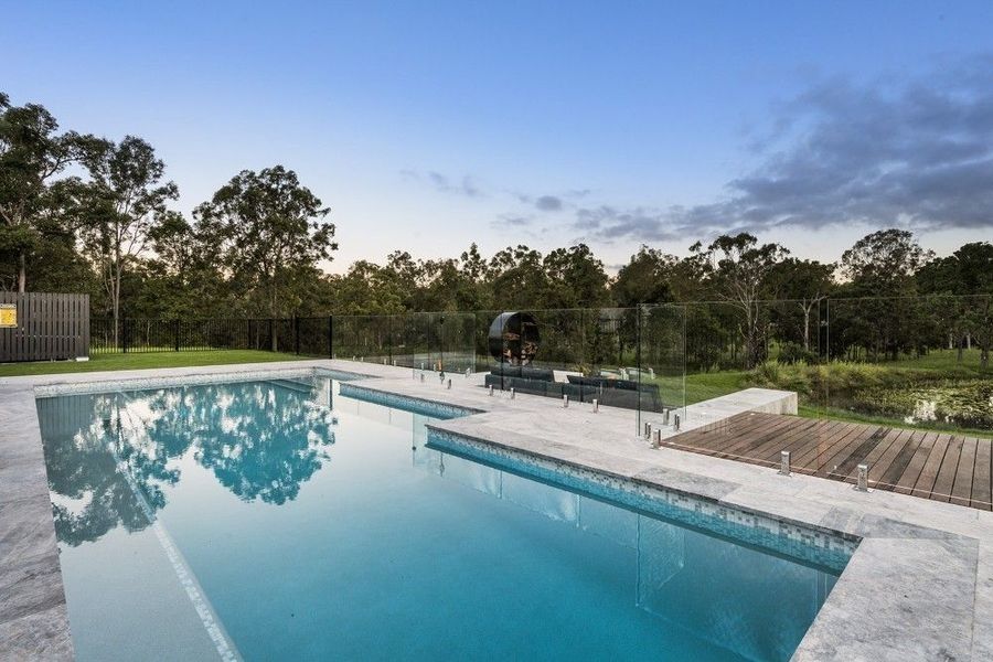 A swimming pool with clear blue water, surrounded by gray stone and a backdrop of trees under a blue sky.