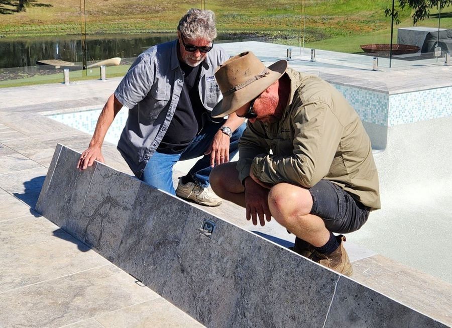 Two men inspecting a long, gray tile near a pool. One kneels, the other stands. Sunny outdoors.