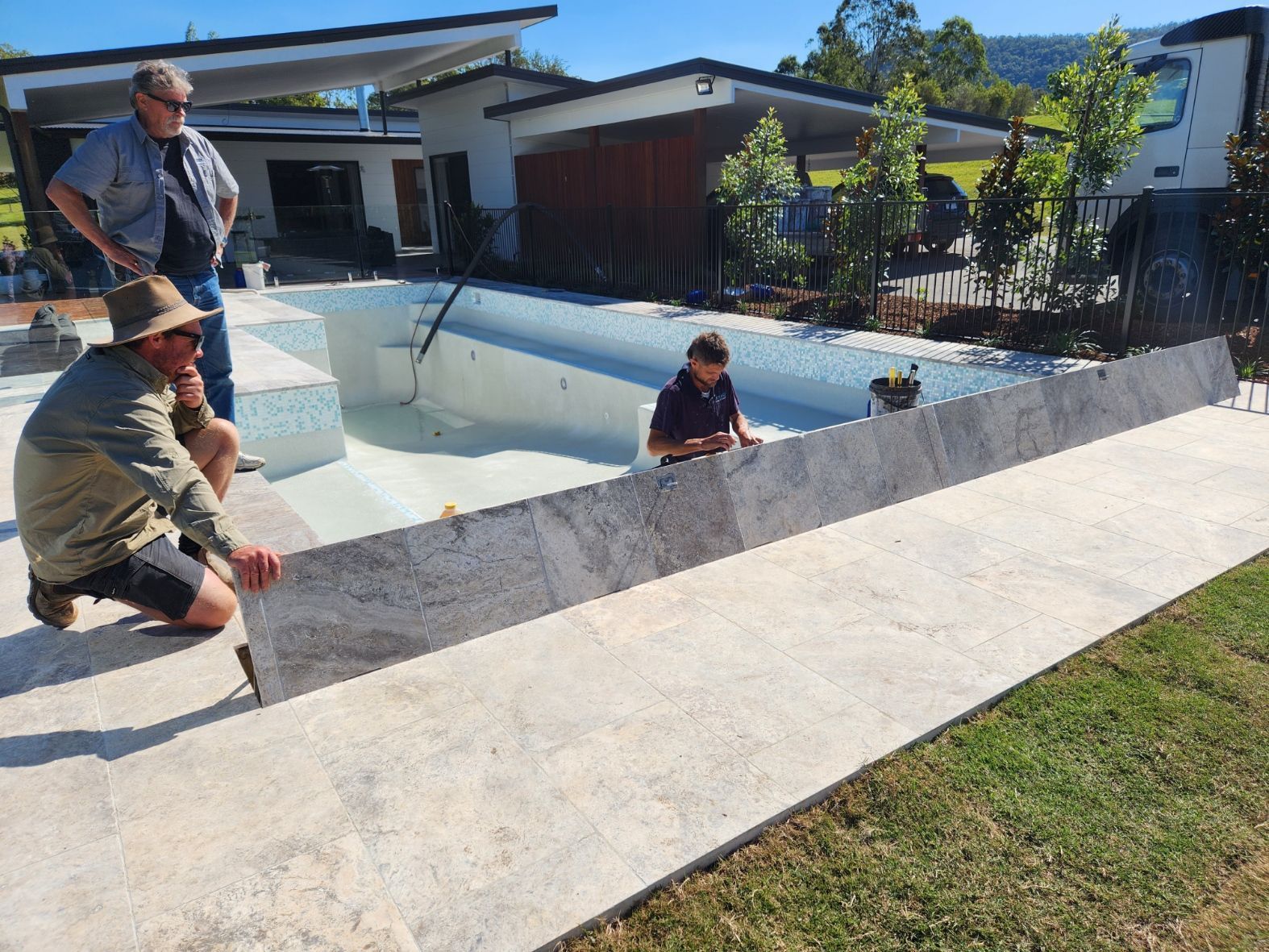 Three people installing gray tile around a pool. Sunlight on a suburban yard.
