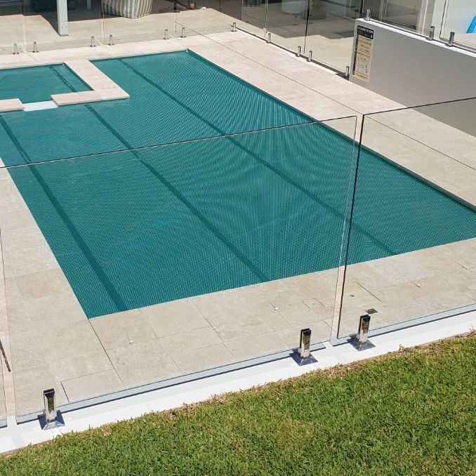 Pool with blue-green water, surrounded by light-colored paving and a clear glass fence, with green grass in the foreground.