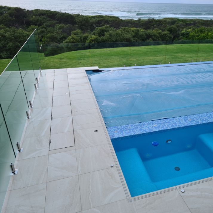 Pool with blue water and cover, glass fence, green grass, and ocean view.
