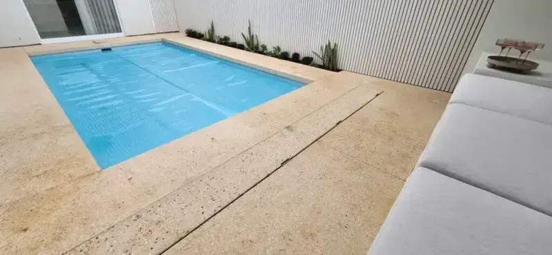 Pool with blue cover, surrounded by tan stone. White wall with plants, and a white couch.