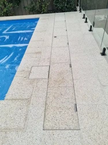 Poolside patio with light-colored speckled tiles. Blue pool cover on left, glass fence on right.