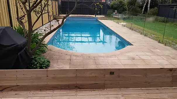 Pool with light blue water surrounded by beige tiled deck and wooden retaining wall, in a yard.