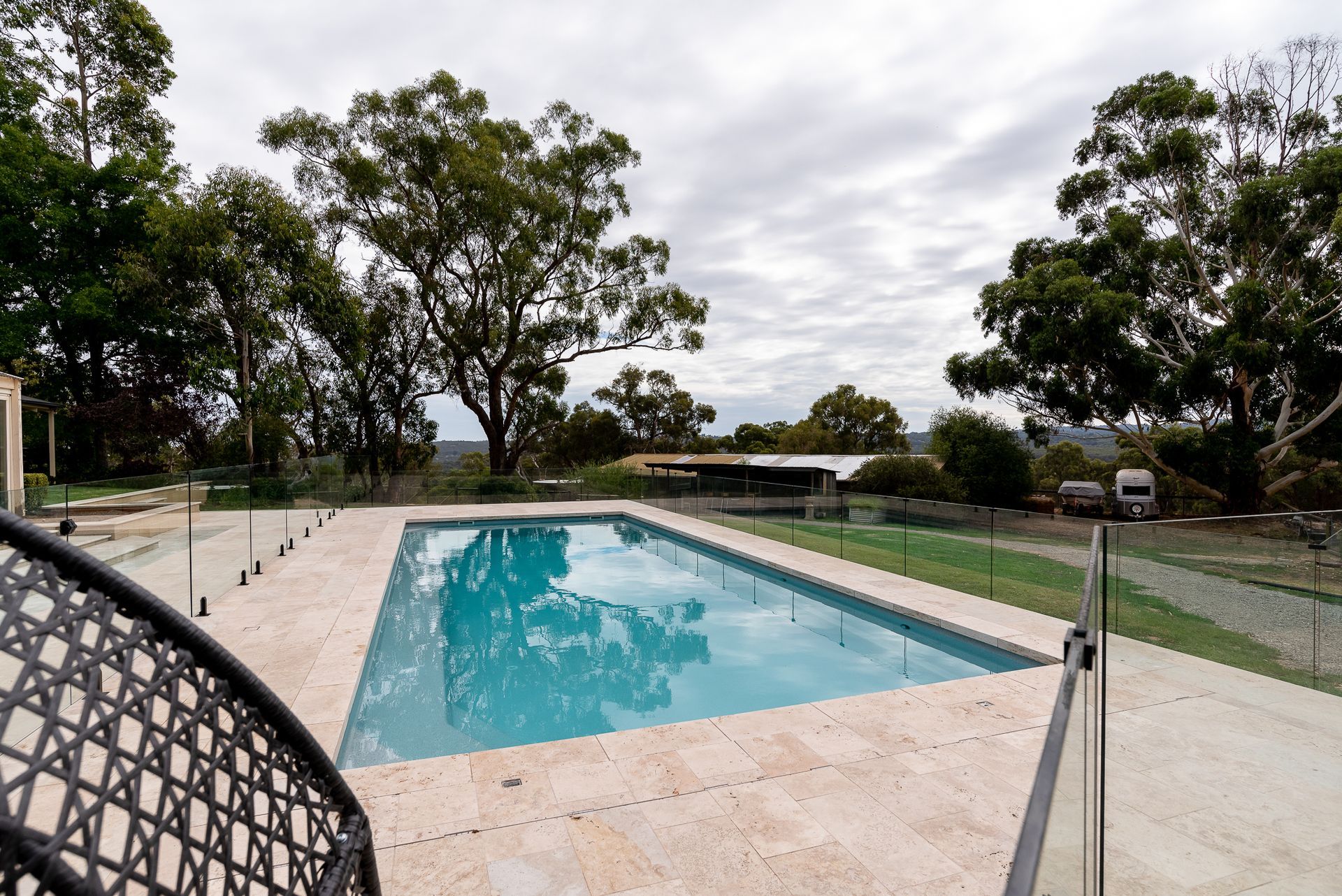 A rectangular swimming pool surrounded by beige tiles and a glass fence, with trees and a cloudy sky in the background.