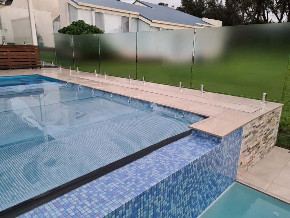Swimming pool with glass fence and frosted glass panels, on lawn with residential houses.