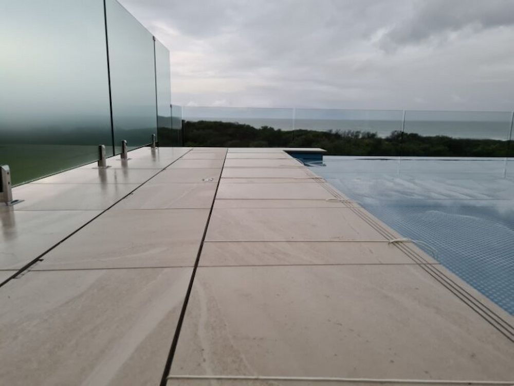 Poolside view overlooking ocean, featuring glass panel railing, tiled deck, and cloudy sky.
