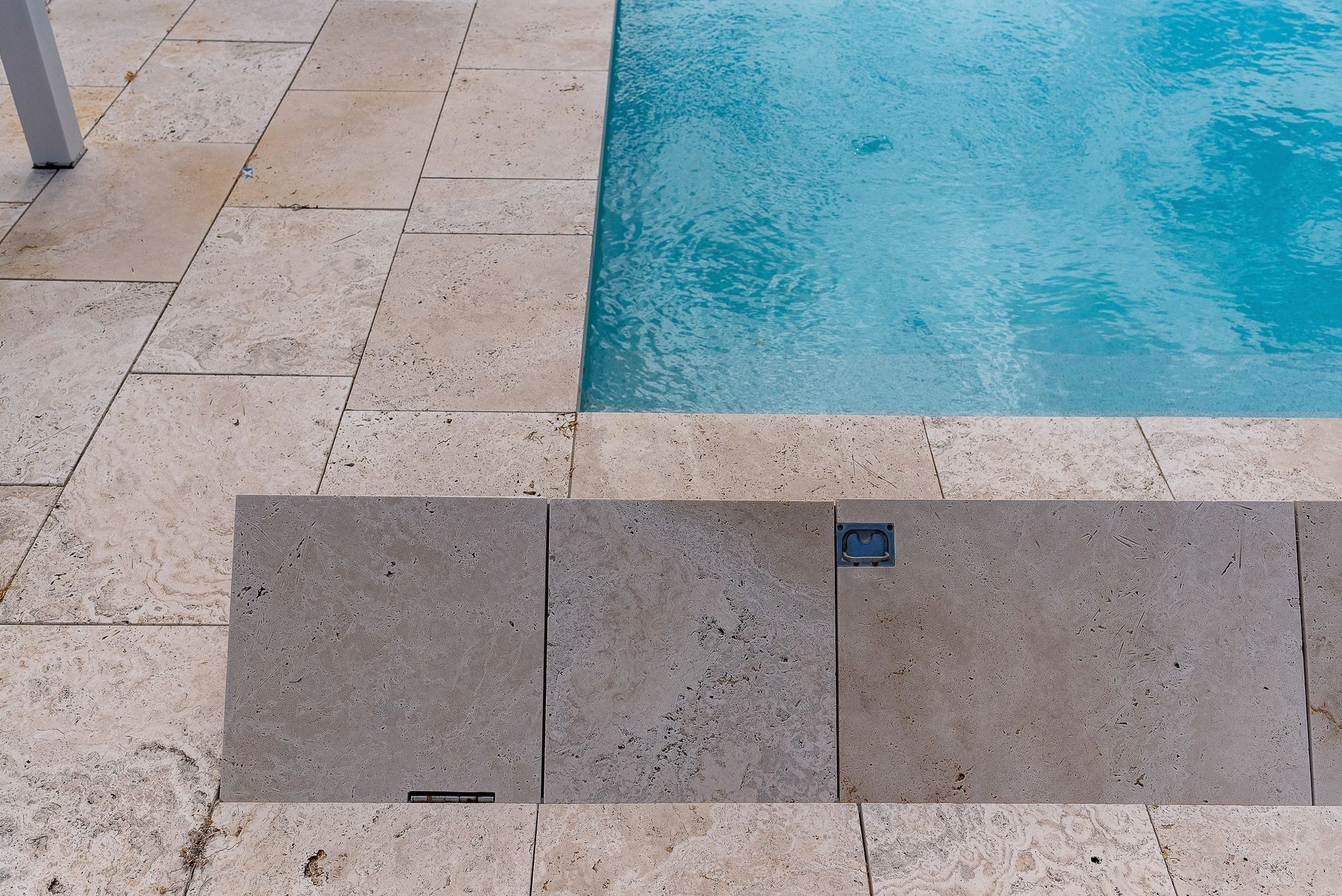 A swimming pool with clear glass fencing. Black posts support the glass, surrounded by greenery and a stone wall.