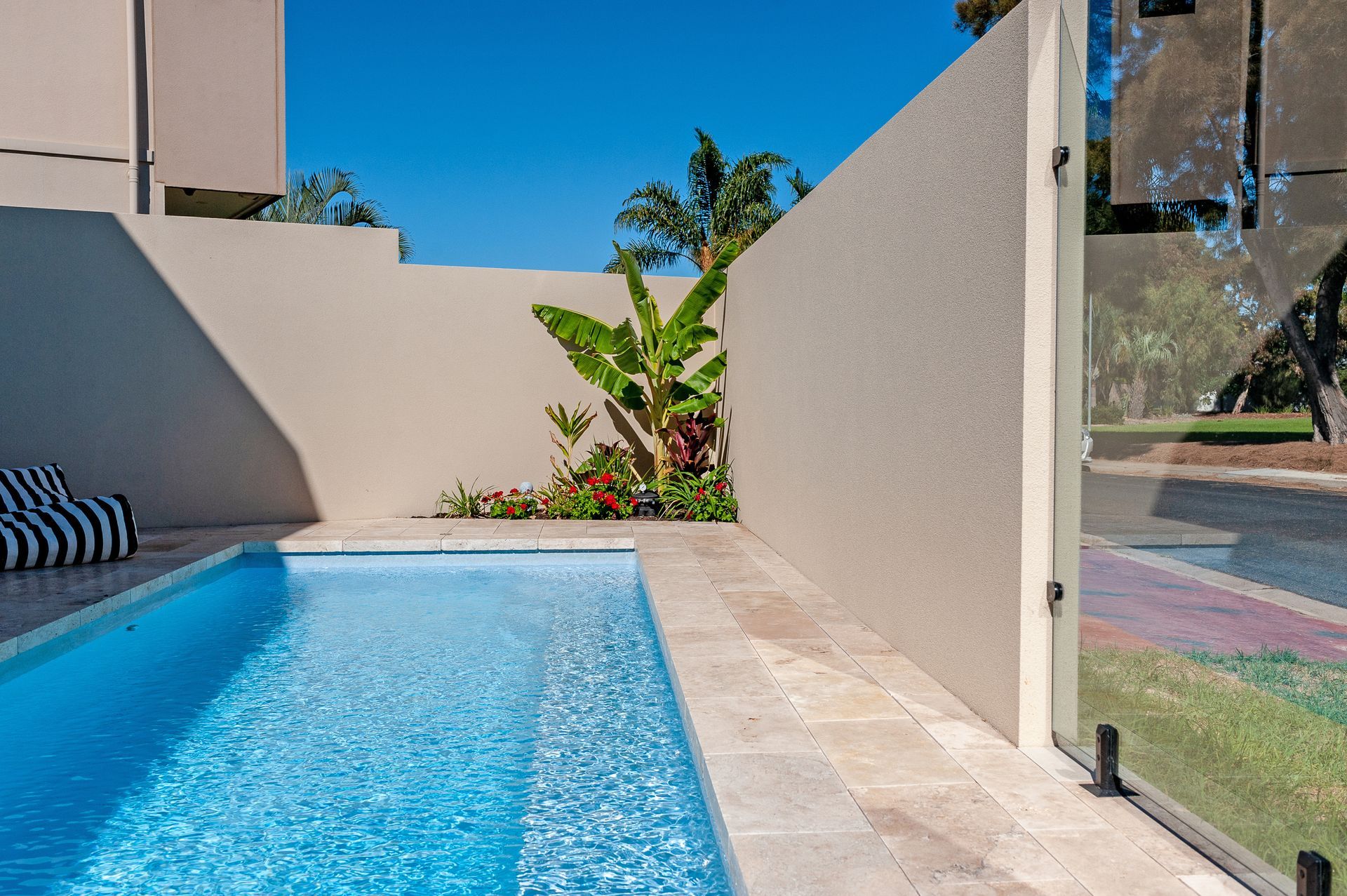 Rectangular blue swimming pool surrounded by stone tiles and a green hedge under a bright sky.