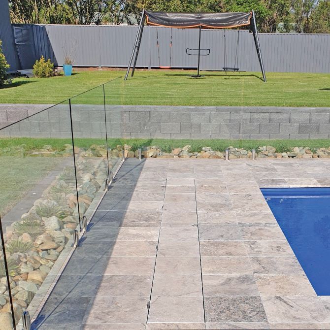 Pool area with glass fence, stone patio, green lawn, swing set, and gray fence.