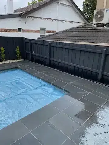 Pool area with blue pool cover, dark grey tile, black fence, and white house in the background.