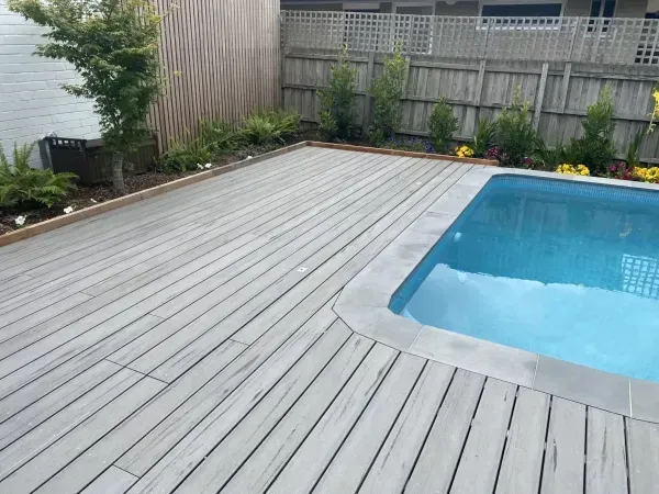 Poolside deck with gray wooden planks, surrounded by landscaping and a pool with blue water.