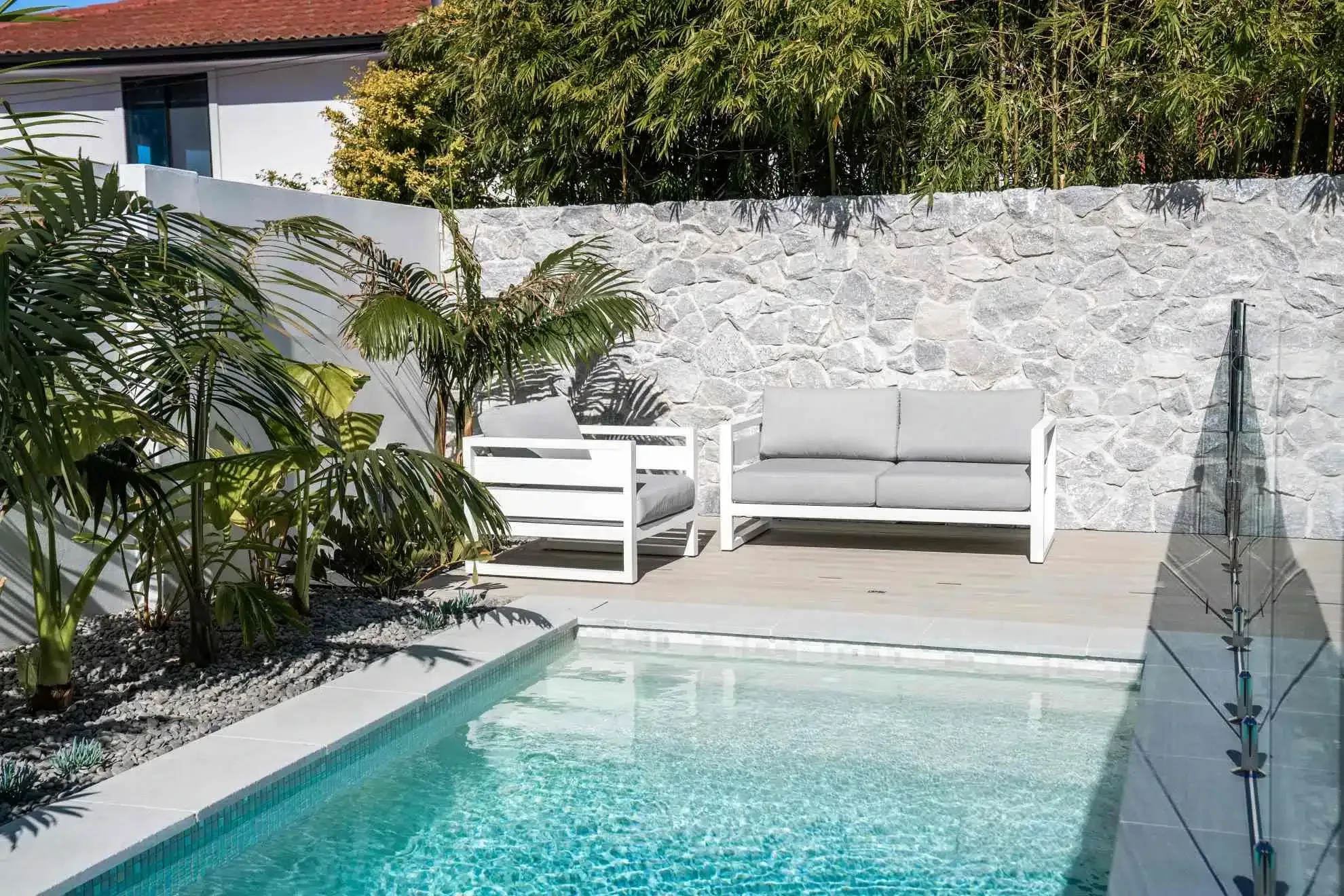 Poolside lounge area with white furniture, stone wall, and tropical plants.