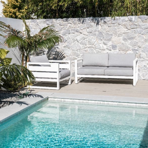 Outdoor patio furniture set next to a pool. White frames, gray cushions. Stone wall backdrop.