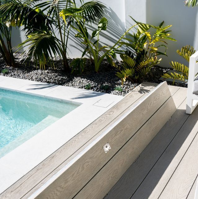 Poolside view with a light gray composite deck and white pool edging; tropical plants in background.