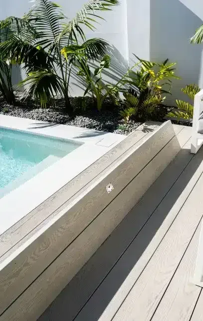 Poolside view: blue pool, light wood decking, stone steps, tropical plants, and white wall.
