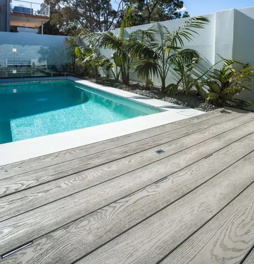 Poolside deck with grey wood planks next to a turquoise pool, white wall, and tropical plants.