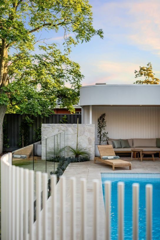 Poolside lounge area with pool and white vertical fence. Teak furniture, stone wall, and lush greenery.