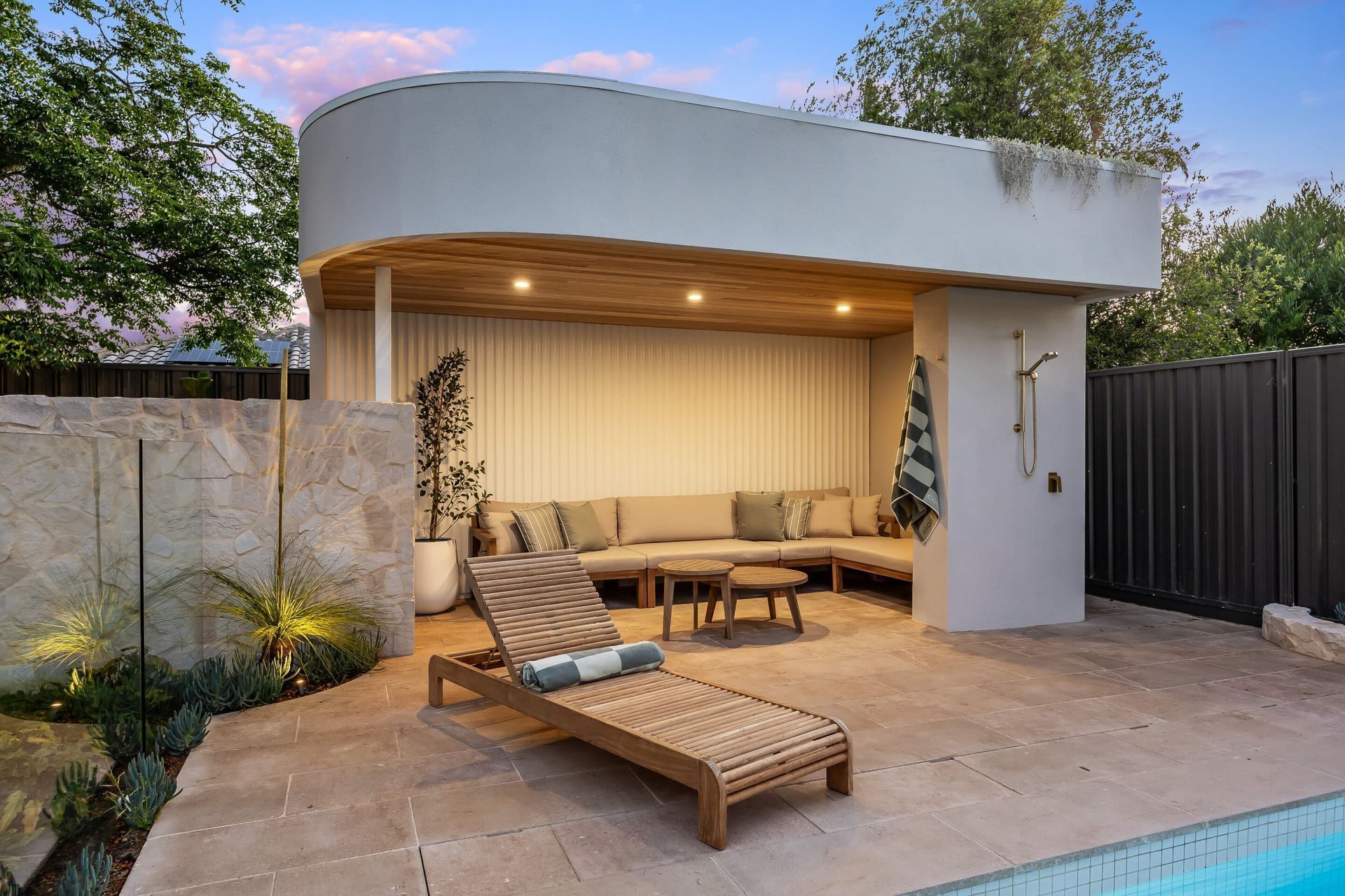 Outdoor lounge area with curved white roof, wooden accents, and a chaise lounge near a pool.
