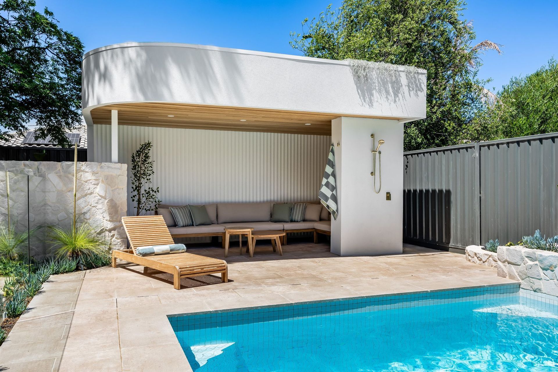 Poolside cabana with lounge seating, wooden chaise, and outdoor shower. Blue pool with turquoise tiles.