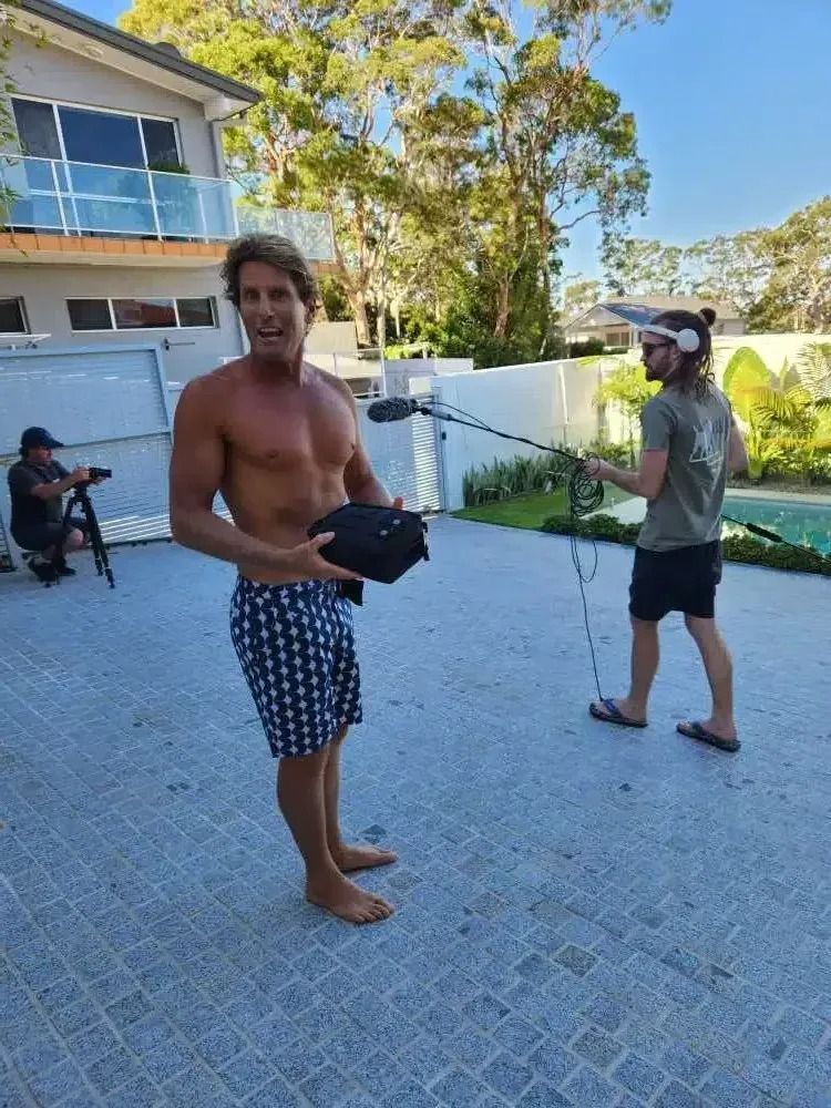 Man holding sound equipment, filming outside a house; crew members in background.