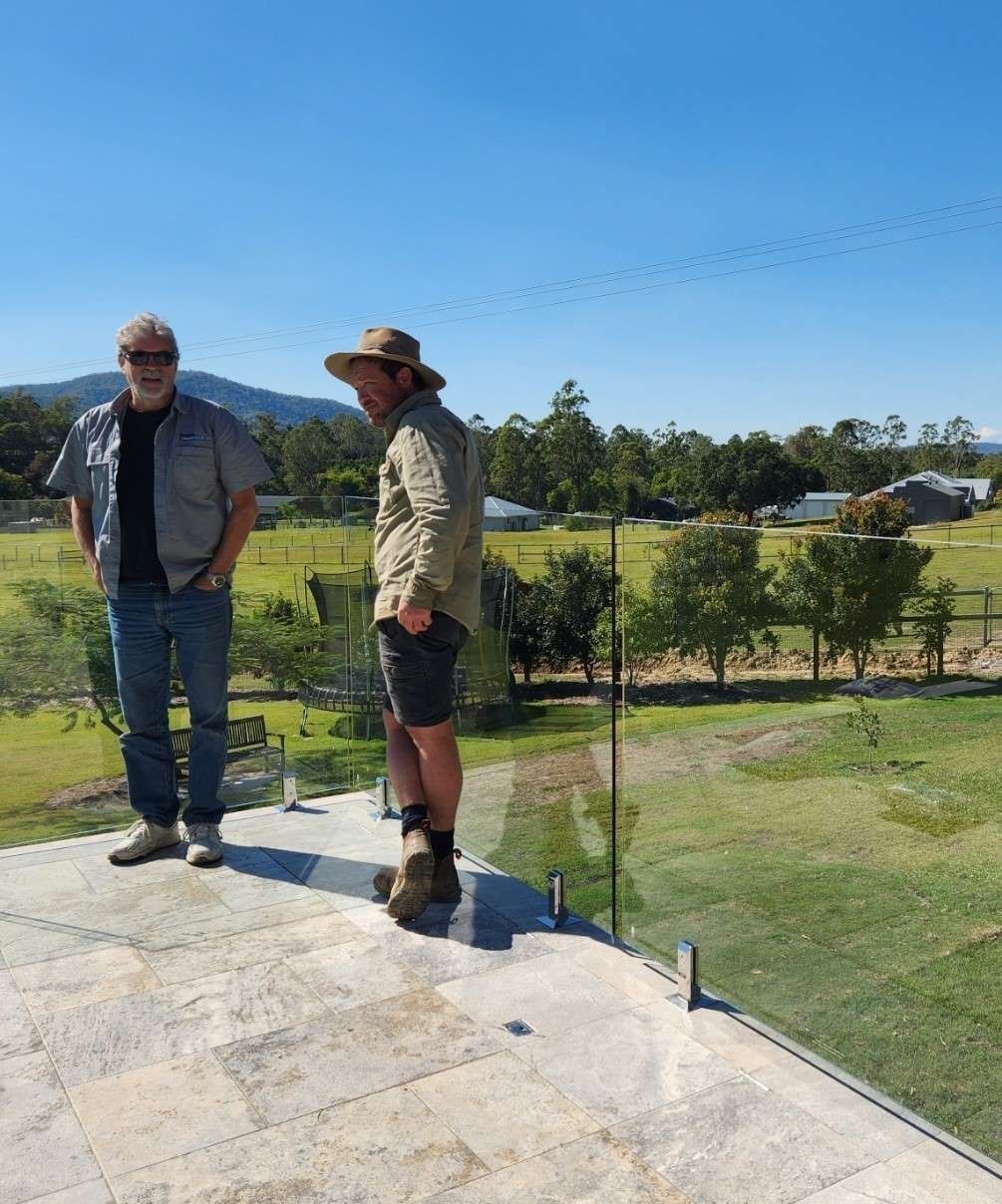 Two men on a tiled rooftop with glass railing, overlooking a green landscape on a sunny day.