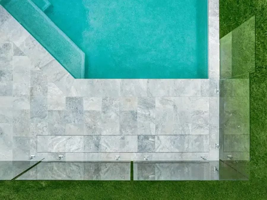 Overhead view of a swimming pool with light blue water, gray stone patio, and glass fencing on green grass.