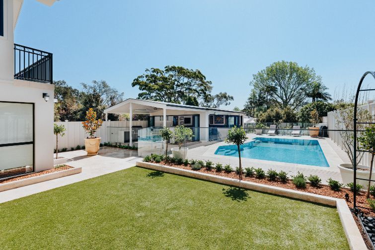 Backyard with pool, patio, and lawn on a sunny day. White house with a balcony and a cabana.
