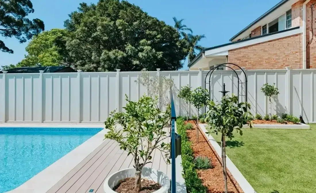 Swimming pool next to a backyard with a white fence, trees, and a brick house.