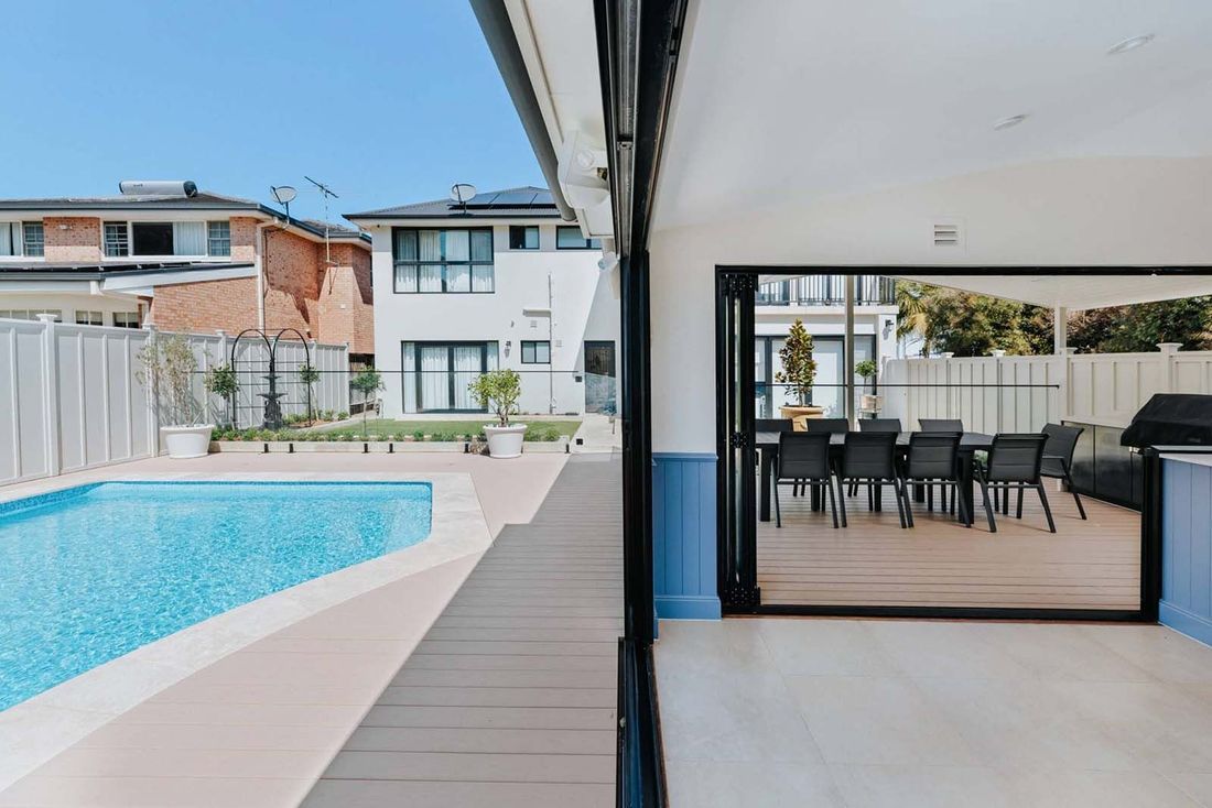 Poolside view with open doors to dining area, blue pool, light-colored deck, and adjacent house.