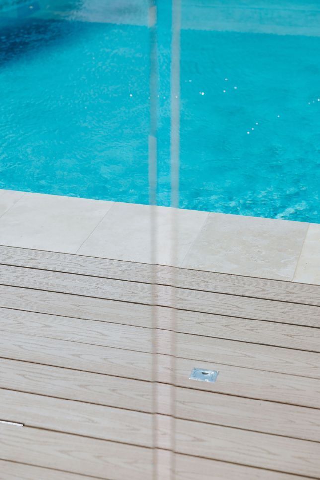 Poolside view with clear water, beige deck, and a light-colored stone border.