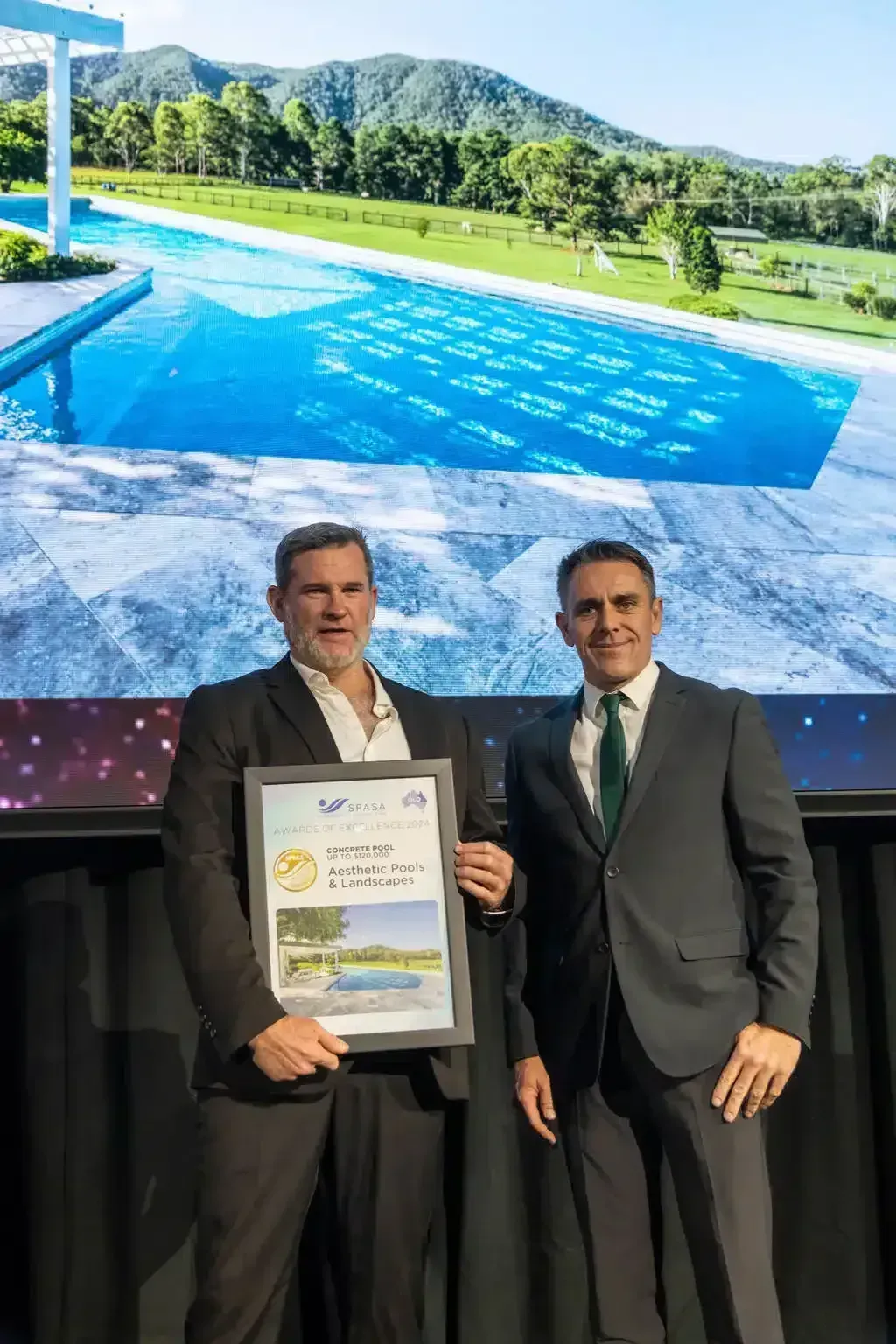 Two men holding award, background features a pool and landscape.
