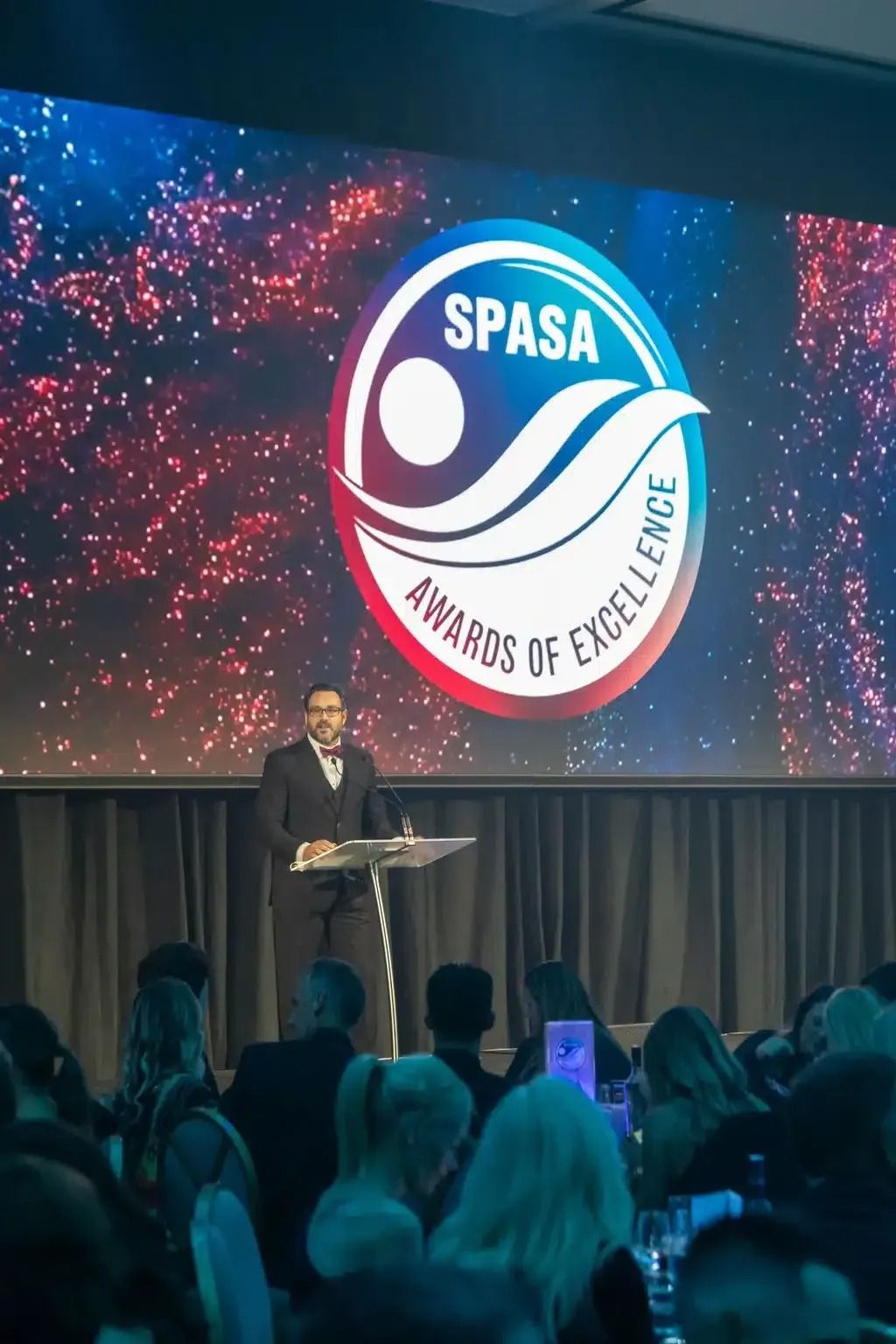 Man speaking at a podium in front of a SPASA Awards of Excellence logo at an event.