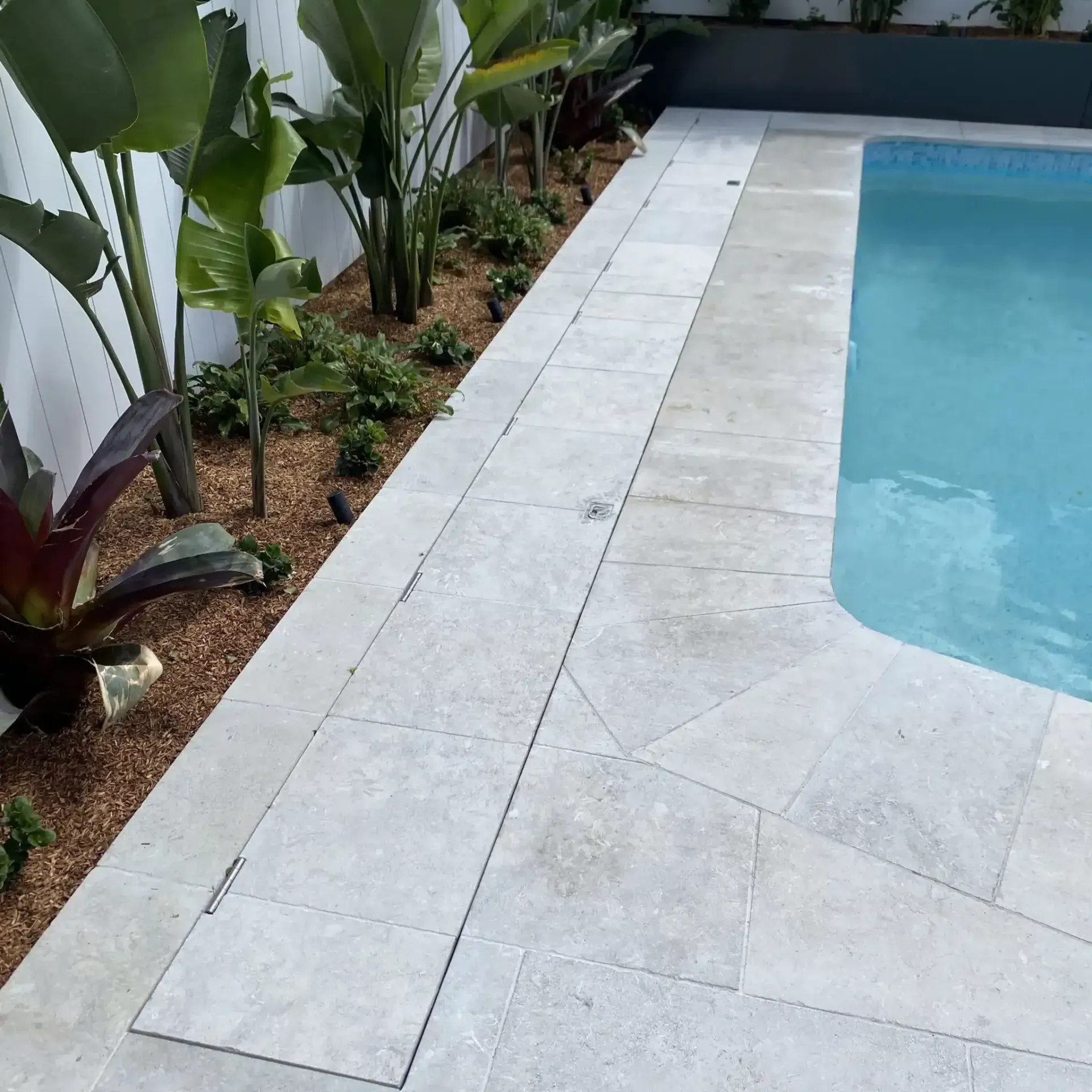 Poolside view with stone tiling, garden bed, and blue water.