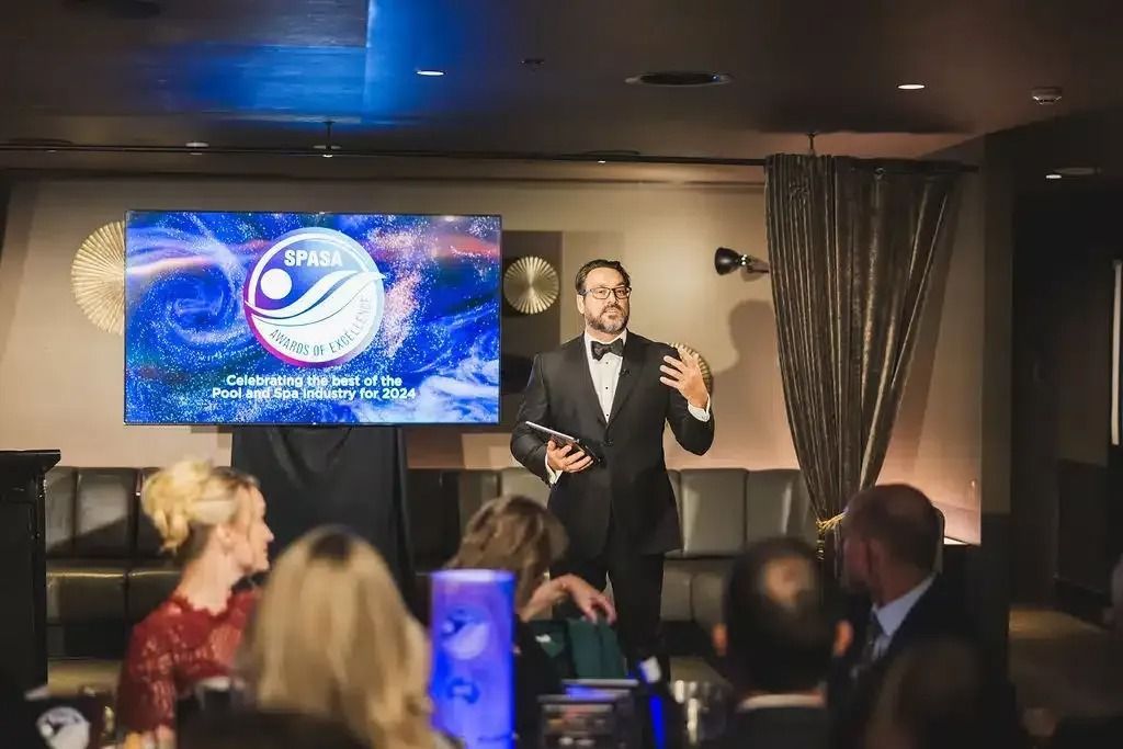 Man in tuxedo speaks at event, logo on screen behind. People seated at tables.