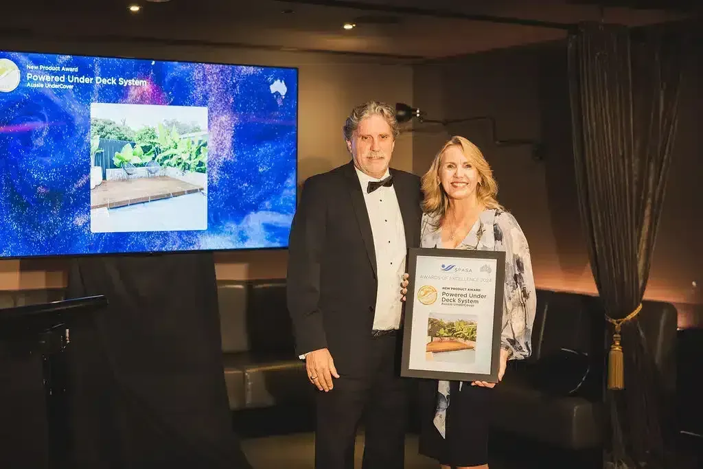 Man in tuxedo and woman holding a framed award, standing in front of a screen displaying a landscape.