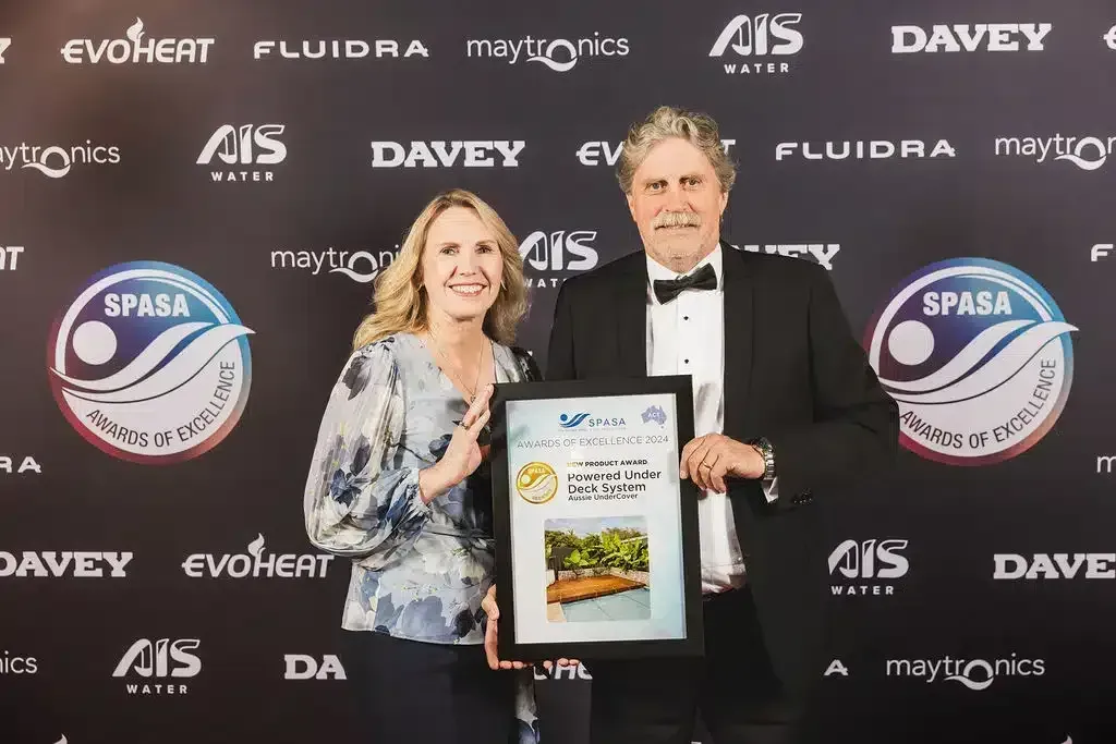 Two people holding an award, smiling at a gala, with sponsor logos in the background.