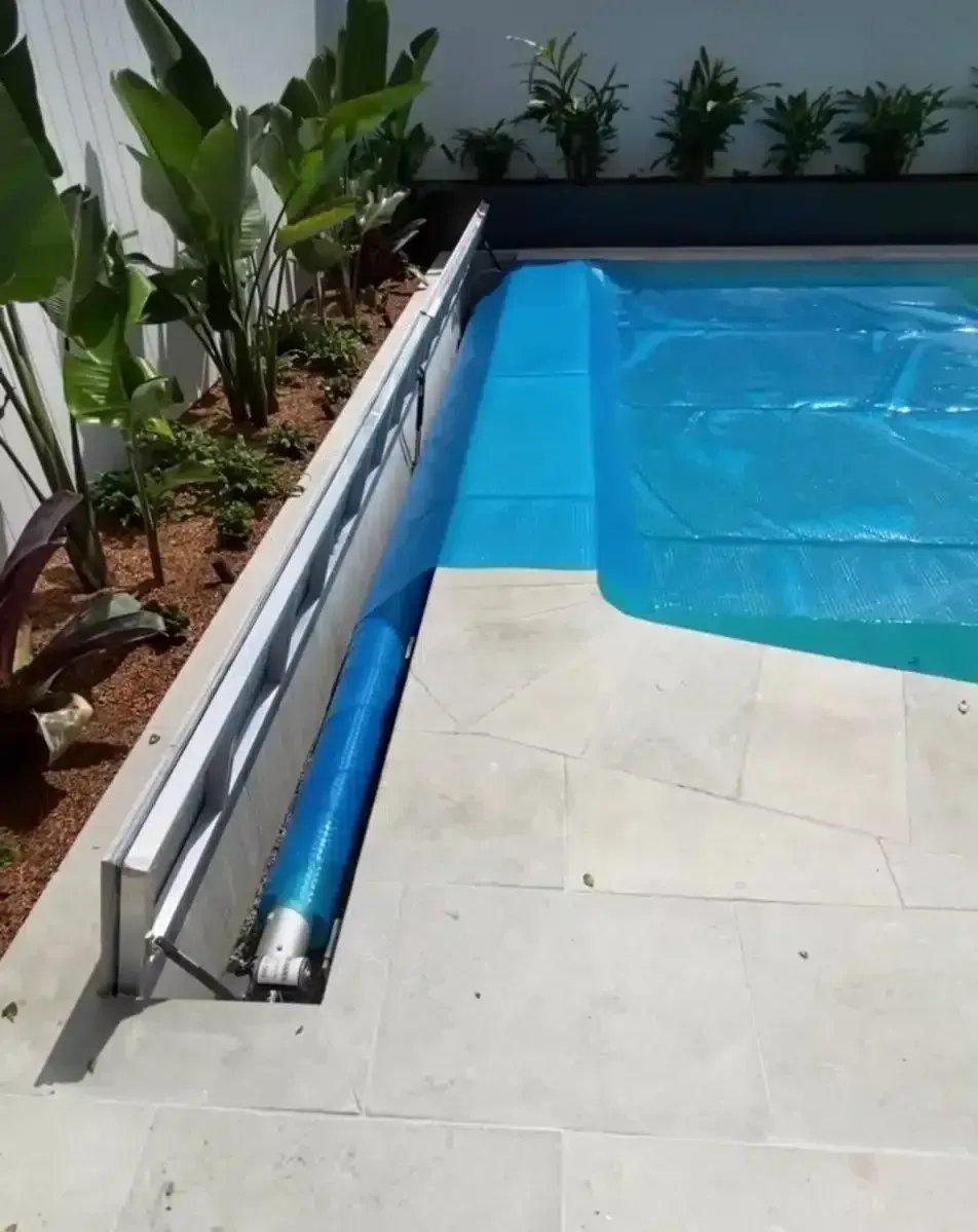 Pool with blue cover partially retracted, revealing rolled-up cover and concrete surround. Lush green plants in background.