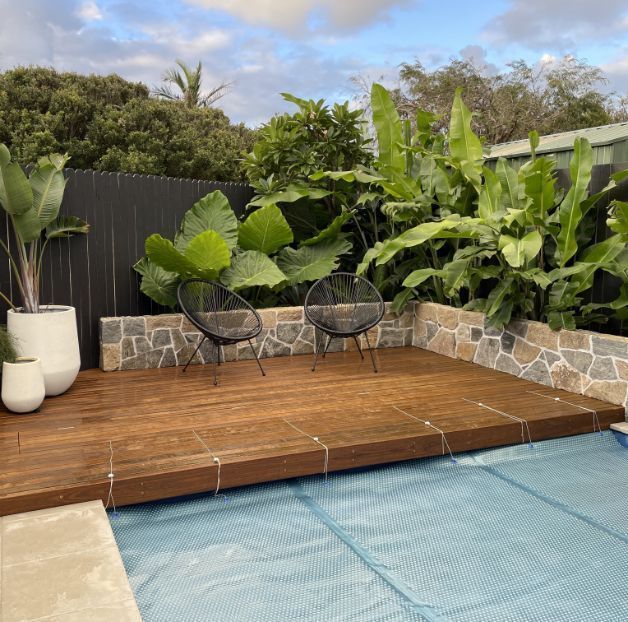 Wooden pool deck with two black chairs, surrounded by lush green plants and stone wall.