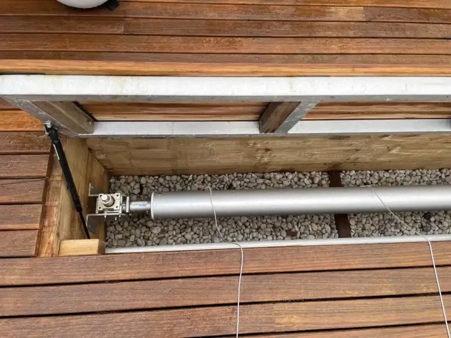 Underside of a wooden deck reveals a silver roller mechanism and gravel.