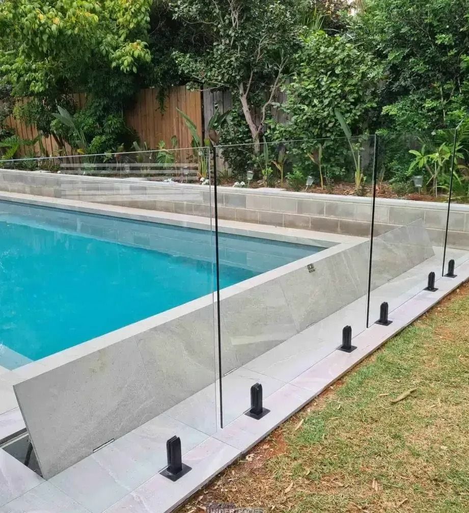 Pool with glass fencing and black posts; set outdoors with green foliage.
