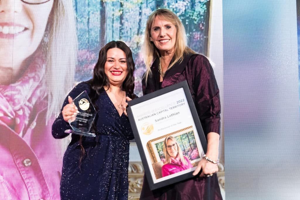 Two women smiling, one holding an award and the other a framed certificate, both on a stage with a screen background.