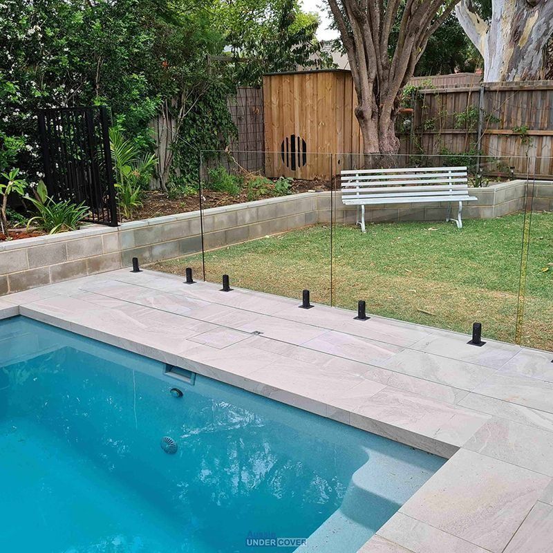 Glass pool fence around a rectangular pool, surrounded by stone and greenery.