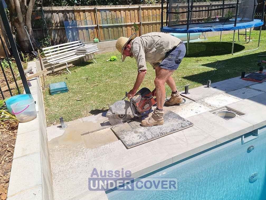 Man cutting a tile next to a pool. Outdoors, sunny. Poolside.