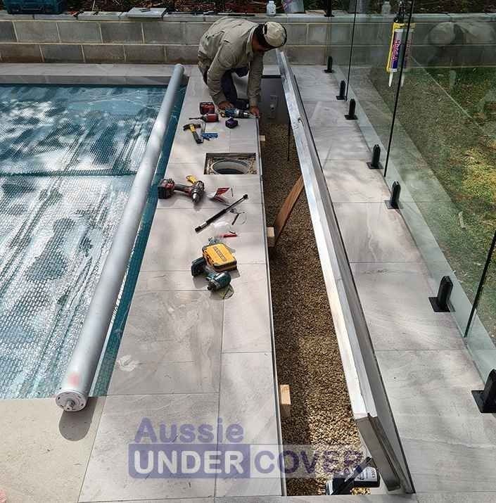 Man installs pool cover track, tools and cover components on poolside tiles.