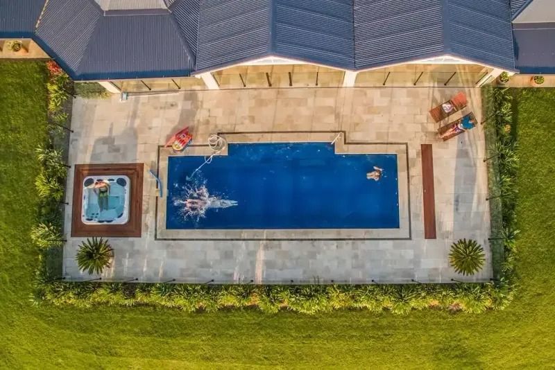 Overhead view of a rectangular pool with people, a jacuzzi, and a house with a dark roof on green grass.