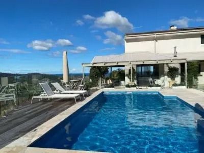 Swimming pool in front of a house, with lounge chairs on a wooden deck. Blue sky with clouds.